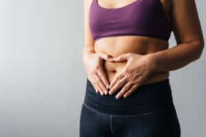 Woman in sports bra and jeans holding hands in heart shape over her tummy after tummy tuck to heal diastasis recti