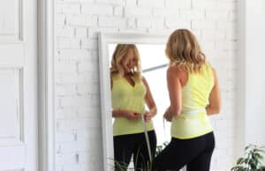 Woman measuring her waist in front of a mirror, showing confidence after a mommy makeover.